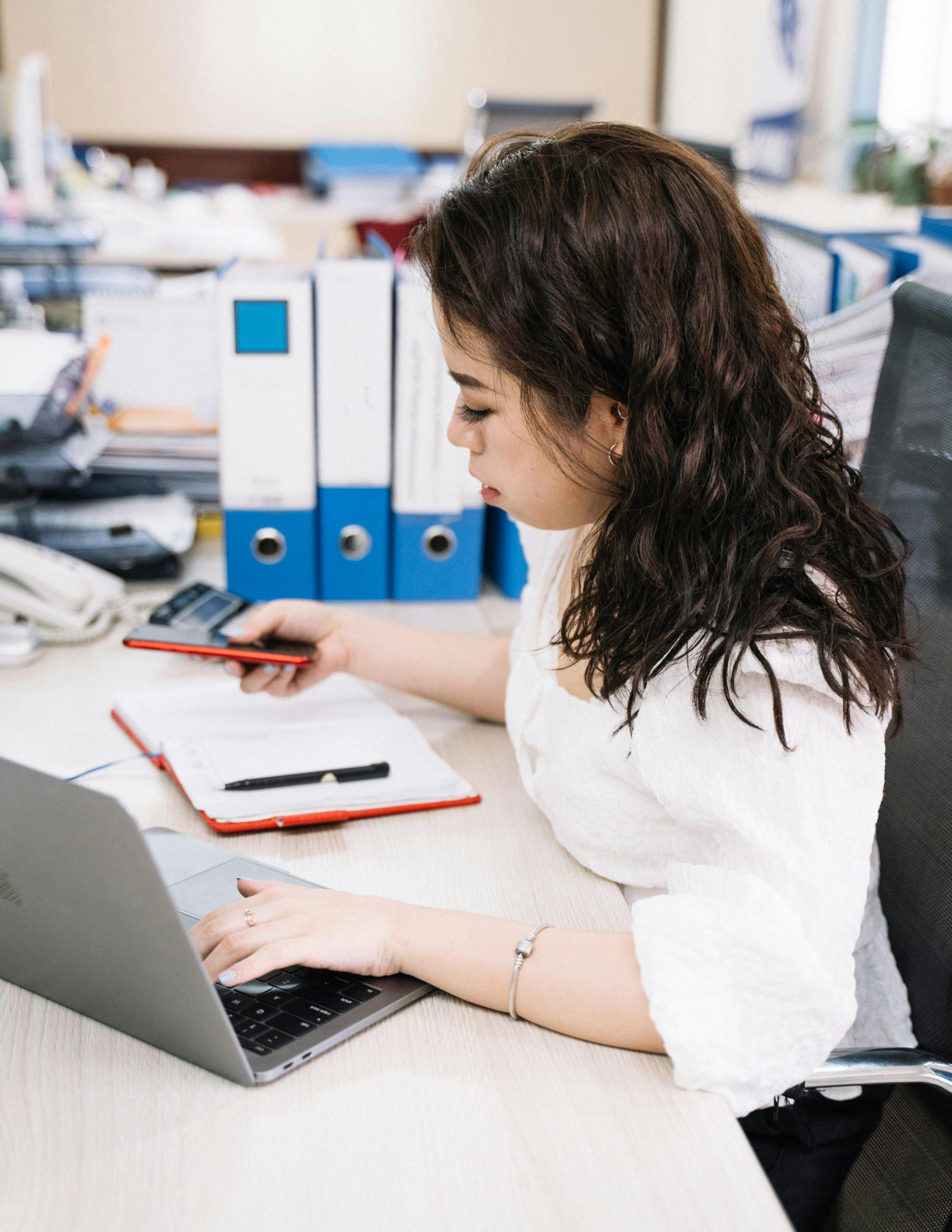 Businesswoman multitasking with phone and laptop at office desk, emphasizing productivity.